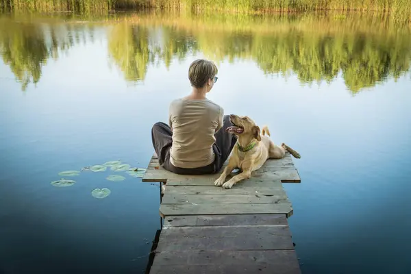 Female with a labrador dog on a wooden fishing bridge enjoying peaceful warm water view. Relaxatioan and spare time with home pets.