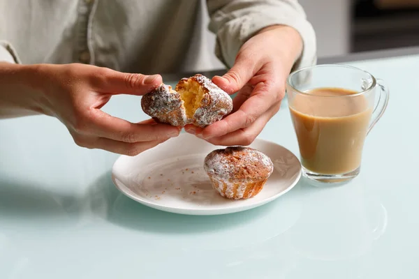 close up of hands breaking apart one of two curd muffins over plate, and glass cup of coffee with milk standing on table Delicious and aroma snack