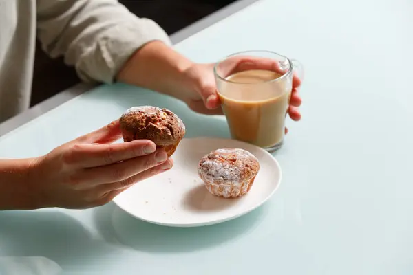 close up of right hand taking one of two curd muffins from plate, and left hand holding glass cup of coffee with milk. Delicious and aroma snack