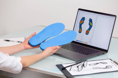 Dişi ortopedist, elinde özel yapım tabanlar bulunan bir klinikte dizüstü bilgisayarın önünde, üzerinde test ayakları resmi olan bir kadın ortopedist. Ayakların dinlenmesi ve ortotik tıp konsepti. Ayak dinlendirme ilacı konsepti