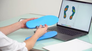 Dişi ortopedist, elinde özel yapım tabanlar bulunan bir klinikte dizüstü bilgisayarın önünde, üzerinde test ayakları resmi olan bir kadın ortopedist. Ayakların dinlenmesi ve ortotik tıp konsepti