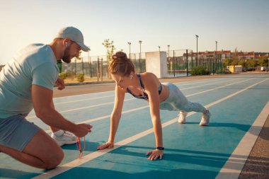 The athletics coach marks the time in the plank exercise on a stopwatch. Fitness trainer and mentee. Instructor and athlete runner on the track.