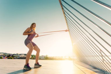 Açık havada trx fitness kayışlarıyla şınav çeken bir kadın. Spor kıyafetli, zinde bir kadın gökyüzünde poz veriyor.