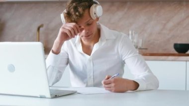 Young adult watching video course and making notes. Student wearing headphones using laptop on his kitchen for learning from online video course and writing down information. Handheld shot