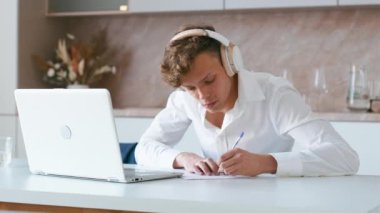 Young adult watching video course and making notes. Student wearing headphones using laptop on his kitchen for learning from online video course and writing down information. Handheld shot