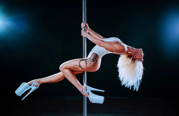 Young blond woman pole dancing on black background with lights. Tattoo on body.