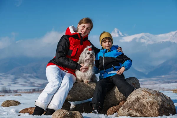 Mother, son and shih tzu dog sitting on stones on winter mountains background