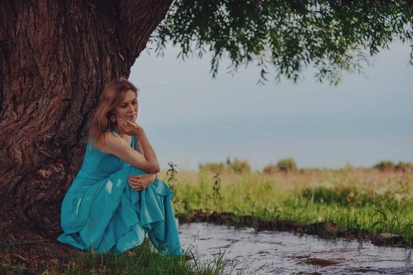 ストリームによって木の下に座って青いドレスの思考エレガントな女性 — ストック写真