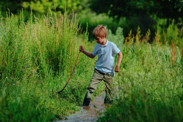 穿着橡胶靴 拿着手杖的男孩沿着一条浅浅的小河走着 — 图库照片