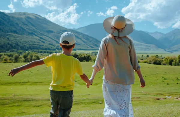 Happy mom and son walking through a green meadow in the mountains holding hands