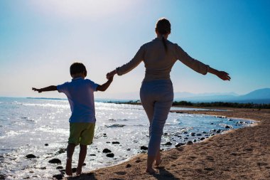 Genç bir kadın ve oğlu kumsalda el ele tutuşarak çıplak ayakla yürüyor.