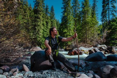 Genç adam nehrin yakınındaki dağ geçidinde büyük bir kayanın üzerinde oturuyor ve başka tarafa bakıyor.