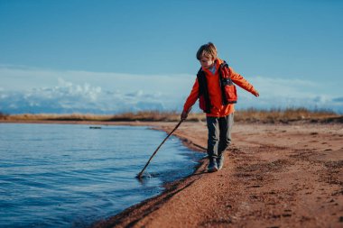 Child tourist with stick walking on lake shore on summer day clipart