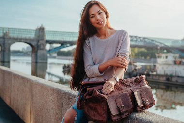 Yaz günü şehirde taştan çitlere oturan çantalı genç bir kadın.