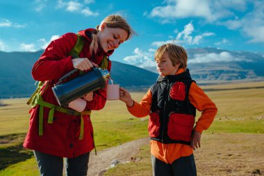 Anne termostan oğlu için dağların arka planında bir fincana çay dolduruyor, turist konsepti.