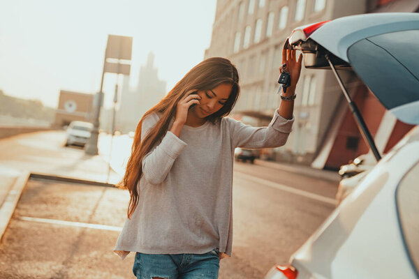 Молодая азиатка открывает дверь багажника машины и разговаривает по телефону летом утром