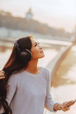 Genç gülümseyen Asyalı kadın şehirde akıllı telefon portresinden müzik dinliyor.