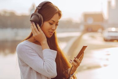 Genç Asyalı kadın nehir kenarında duruyor ve akıllı telefondan müzik dinliyor.