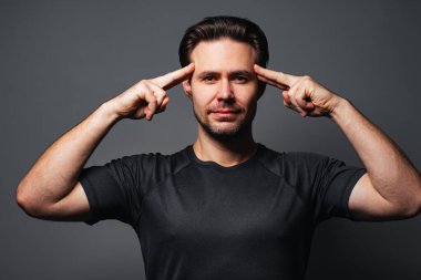 Genç yakışıklı adam parmaklarıyla şakaklarına dokunuyor, gri arka planda portresi var.