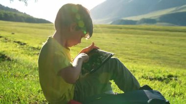 Çocuk dağlarda çayırda oturuyor ve tablete bakıyor.
