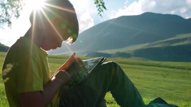 Çocuk dağlarda çayırda oturuyor ve tablete bakıyor.