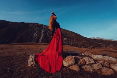 Genç, güzel, kırmızı elbiseli bir dansçı akşam yüksek dağlarda dikiliyor.