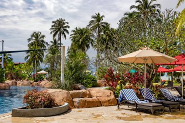 Malezya, Langkawi, Kasım 2022: Şemsiyelerin altında güneşlenen şezlonglar, Langkawi 'deki Pelangi Beach Spa Oteli' nin bölgesinde uzun palmiye ağaçları olan güzel bir bahçenin içinde yüzme havuzunun yanında.