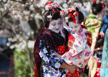 Kollarında küçük bir kız olan hoş bir bayan. İki Maiko, şef ve küçük, çırak Geyşa, güzel bir kimono giyiyor, kiraz çiçeklerinin arka planında. Gion, Kyoto.