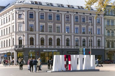 LATVIA, RIGA, MAYIS, 2023: Riga Old Town 'daki McDonalds Kafe arka planında Letonya' nın adını taşıyan büyük harflerle yürüyen insanlar. Riga şehir tarihi merkezi UNESCO 'nun Dünya Mirasları Alanıdır.