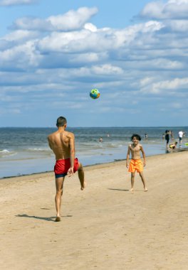 LATVIA, JURMALA, 07, JULY, 2023: Baltık Denizi sahilinde küçük bir çocukla futbol oynayan atletik adam, Jurmala. Spor anlayışı, çocukluk, aktif yaşam tarzı.