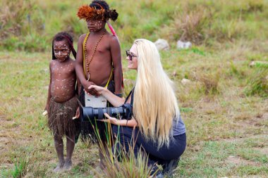 INDONESIA, PAPUA NEW GUINEA, WAMENA, IRIAN JAYA, 20 AUGUST 2018: A woman with long hair and a camera in her hand sits next to and holds the hand of the children from the papuan tribe in Wamena, Papua