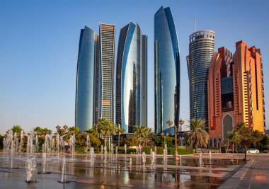 BAE, ABU DHABI, NOVEMBER, 18, 2021 - Birleşik Arap Emirlikleri 'nin başkenti Abu Dabi' de günbatımında yeşil palmiye ağaçları ve çeşmelerle çevrili gökdelenler.