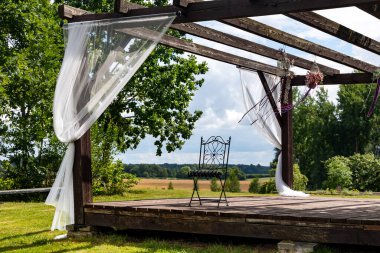 Beyaz şeffaf perdeleri ve bahçe mobilyaları olan ahşap çardak Letonya kırsalında kabarık bulutlu bir gökyüzünün arka planına karşı.