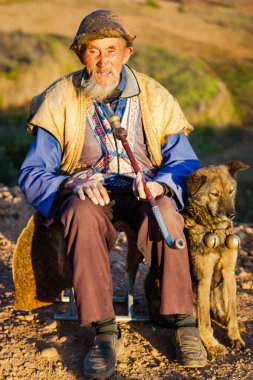 CHINA, DONGCHUAN, YUNNAN PROVINCE, NOVEMBER, 11, 2018 - Elderly and cheerful chinese peasant in national clothes with a long tobacco pipe and your favorite dog in the mountains of Dongchuan, Yunnan clipart