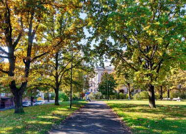 LATVIA, RIGA, 15, ECTOBER, 2024: İnsanlar Riga, Letonya 'da güneşli bir günde sonbahar yapraklarıyla bir parkta bir bankta oturuyor