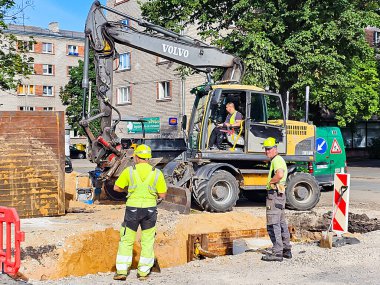 LATVIA, RIGA, AUGust, 21, 2025: İnşaat işçileri Riga, Letonya 'nın merkezindeki yerleşim binaları arasında bir kazıcının çalışmasını izliyor.