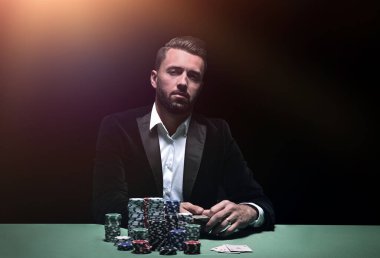 Önünde fişleri ve kartlarıyla poker masasında oturan genç adam.