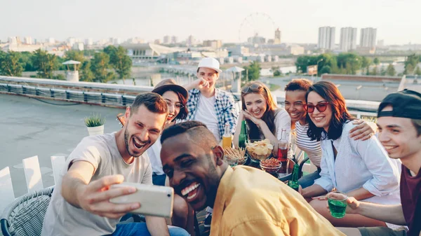 幸せな若い男性と女性は ポーズ 笑って 食べ物や飲み物とテーブルで屋根の上に座っての祝日を祝う手ジェスチャーを作るスマート フォンと Selfie を取っています — ストック写真