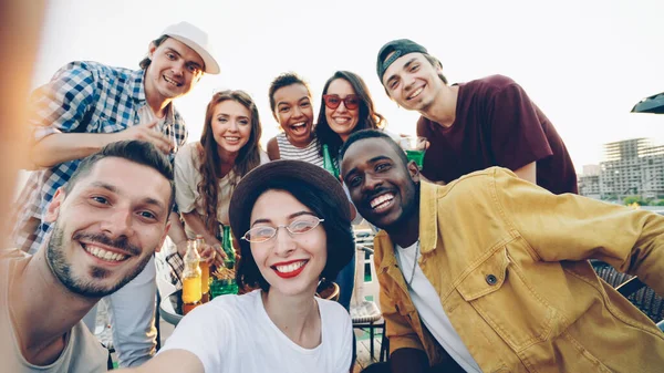 stock image Point of view shot of pretty young lady in trendy hat holding camera and taking selfie with friends enjoying rooftop party with soft drinks, people are looking at camera and laughing.