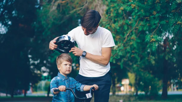 Caring father is putting safety helmet on his little sons head then teaching happy boy to ride bicycle in beautiful park