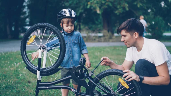 Curious child wearing helmet is spinning bicycle wheel and pedals while his careful father is talking to him on lawn in park on summer day. Family, leisure and active lifestyle concept.