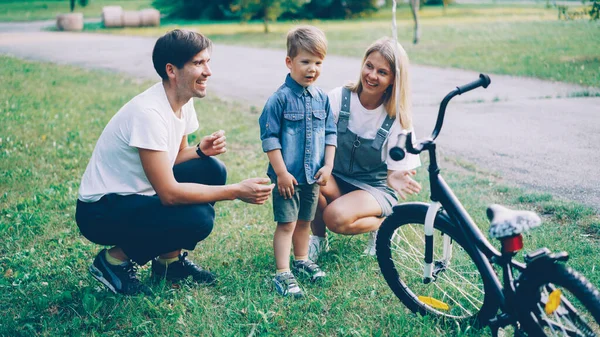 Loving parents are making surprise for little son closing his eyes and giving him new bicycle as present, happy excited boy is looking at bike and talking to mother and father.