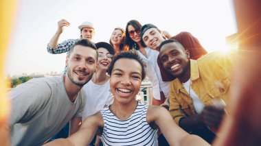 Oldukça Afro-Amerikan Kız holding kamera çekimi selfie çatıda partide mutlu arkadaş grubu bir kadeh bakış açısı. Kadın ve erkek kameraya bakarak, poz ve gülüyor.
