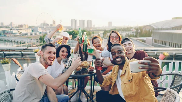 若いアフリカ系アメリカ人男性はスマート フォンを持って ポーズ 屋上パーティーを楽しんで ボトルとグラスを保持している彼の友人の多民族グループを Selfie を取って — ストック写真