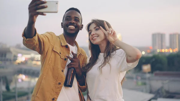 魅力的な混血カップルは屋上パーティーでスマート フォンで Selfie を取っている 男と女を飲み物を保持していると バック グラウンドで夜市と変な顔とポーズ — ストック写真