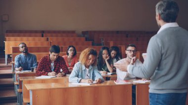 Profesör grup raporları ile sınıfta ayakta ve gençler için onu dinlerken konuşma ve yazma çalışma masaları, oturma çok ırklı öğrenci için ders okuma.