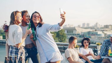 Genç ve güzel bayanlar selfie onların arkadaşları arka planda sohbet çatısında açık hava partisi sırasında smartphone kullanarak şişe ile alıyor. Modern teknoloji ve eğlence kavramı.