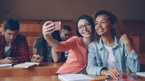 学生は大学でテーブルに座ってのスマート フォン Selfie を取っている元気な女の子 若い女性がポーズをとって手ジェスチャーを作ると抱き締めます 友人や技術コンセプト — ストック写真