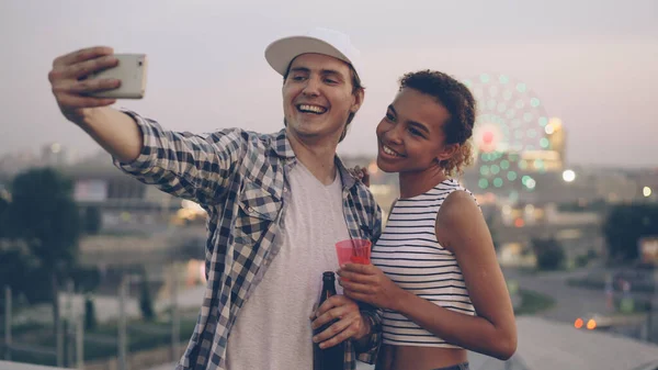 陽気な多民族カップル白人の若い男とアフリカ系アメリカ人の女の子は スマートフォンを使って屋上で自撮りをしています 背景に都市の照明と笑いと飲み物を保持 — ストック写真