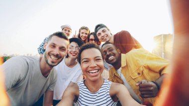 Oldukça Afro-Amerikan Kız holding kamera çekimi selfie çatıda partide mutlu arkadaş grubu bir kadeh bakış açısı. Kadın ve erkek kameraya bakarak, poz ve gülüyor.